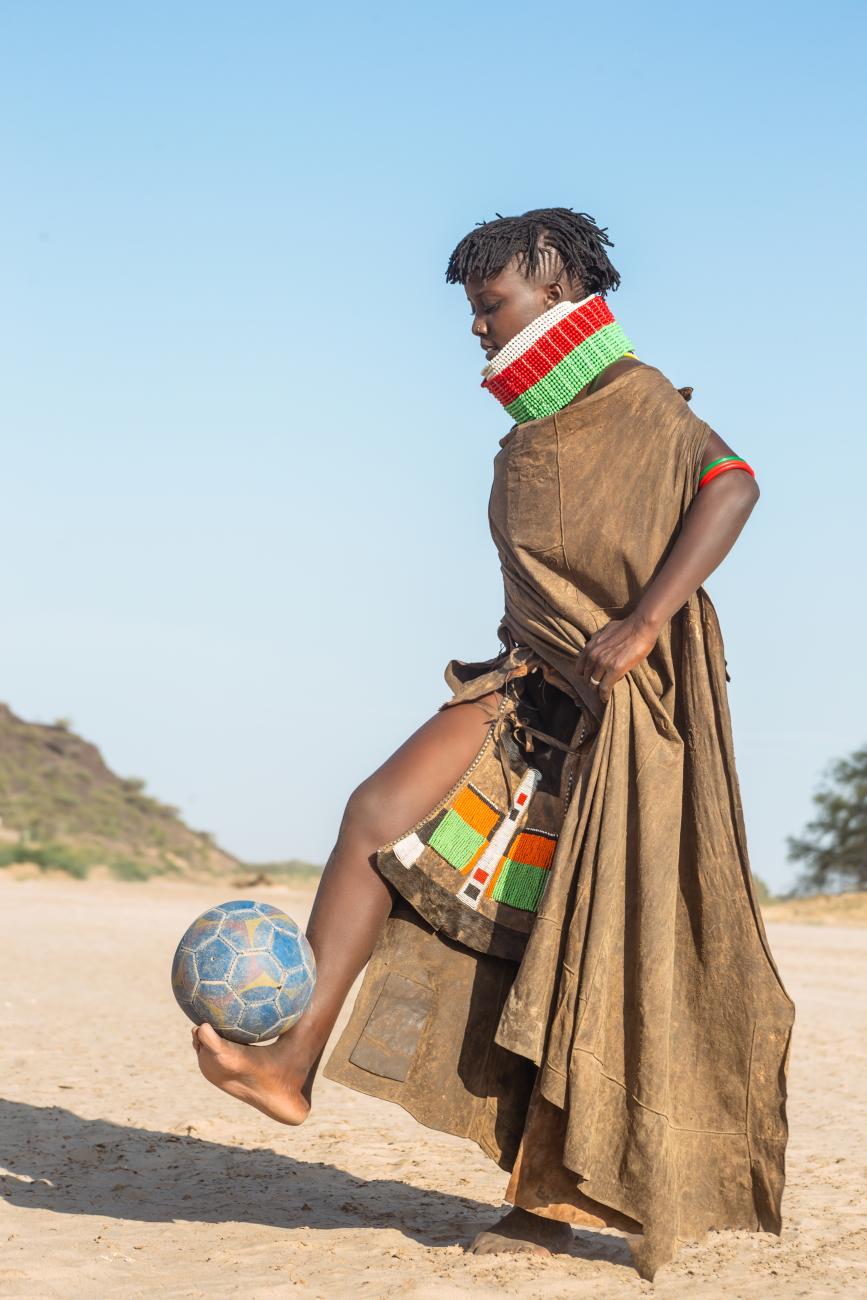 A young woman in traditional attire with colourful beadwork balances a football barefoot on her foot in a sandy landscape.
