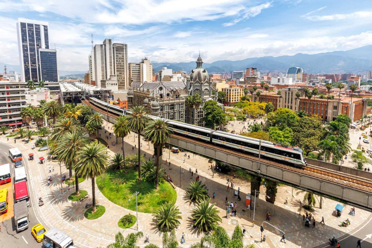A modern train travels on an elevated track through a city with palm trees, historic and tall buildings under a blue sky.
