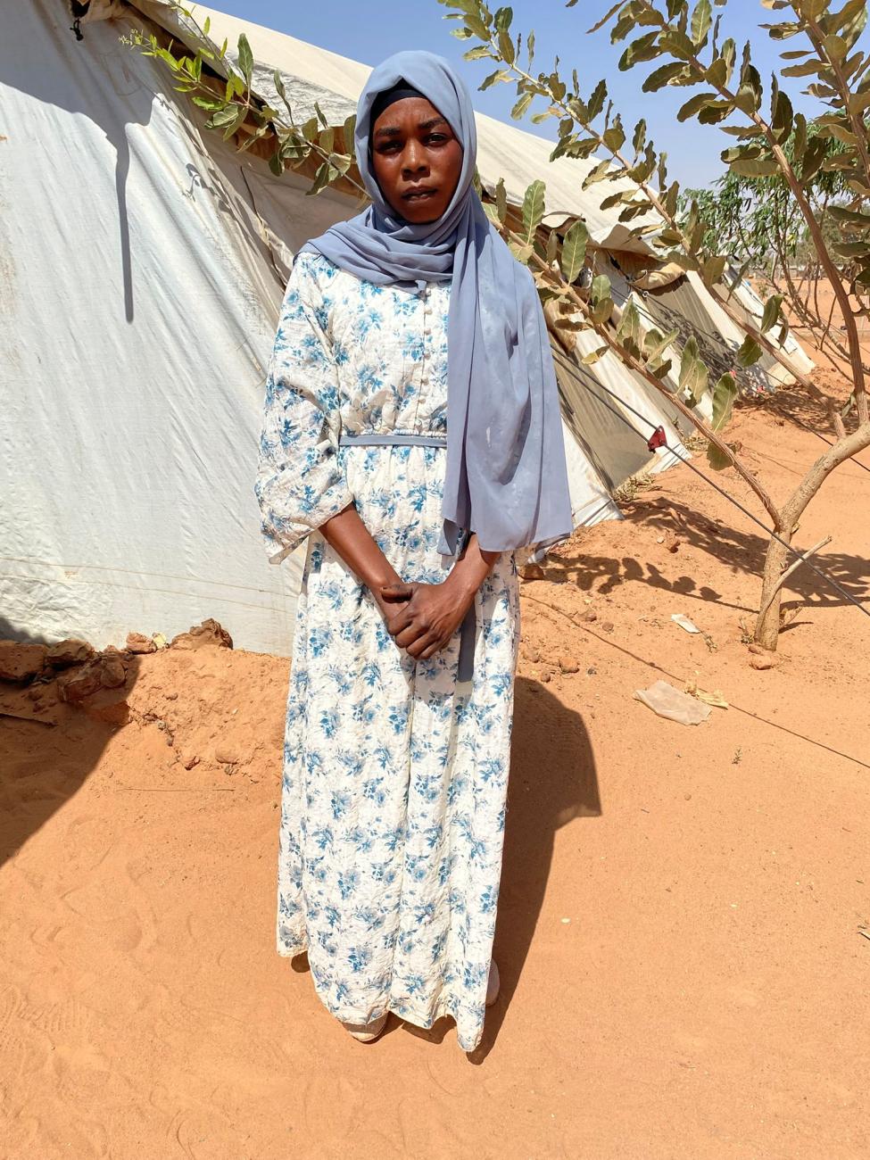 A young woman in a grey headscarf and blue floral dress stands on sandy ground in front of light-coloured tents and bushes.