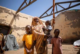 Noory Taha for the IRC Eine Frau in einem gestreiften Gewand hält ein Kleinkind auf dem Arm und steht mit zwei weiteren Kindern vor den Überresten eines zerstörten Hauses mit beschädigtem Dach unter freiem Himmel.