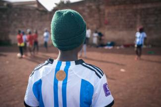 Ein Kind mit grüner Strickmütze und blau-weiß gestreiftem Fußballtrikot steht mit dem Rücken zur Kamera und schaut auf andere Kinder, die auf einem staubigen Platz vor einer Mauer Fußball spielen.